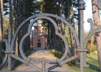 Fotografie des Friedhofseingangs in Gardelegen. Durch ein verziertes Eisengitter ist der Weg zur Kapelle zu sehen, gesäumt von hohen Bäumen.